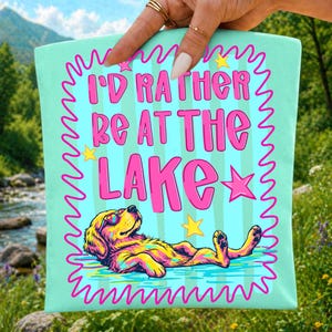 May include: A light mint green fabric pouch featuring a graphic print of a golden retriever wearing sunglasses, lying down in water. The text reads I'D RATHER BE AT THE LAKE in bold pink letters, surrounded by a pink squiggly border and yellow stars.
