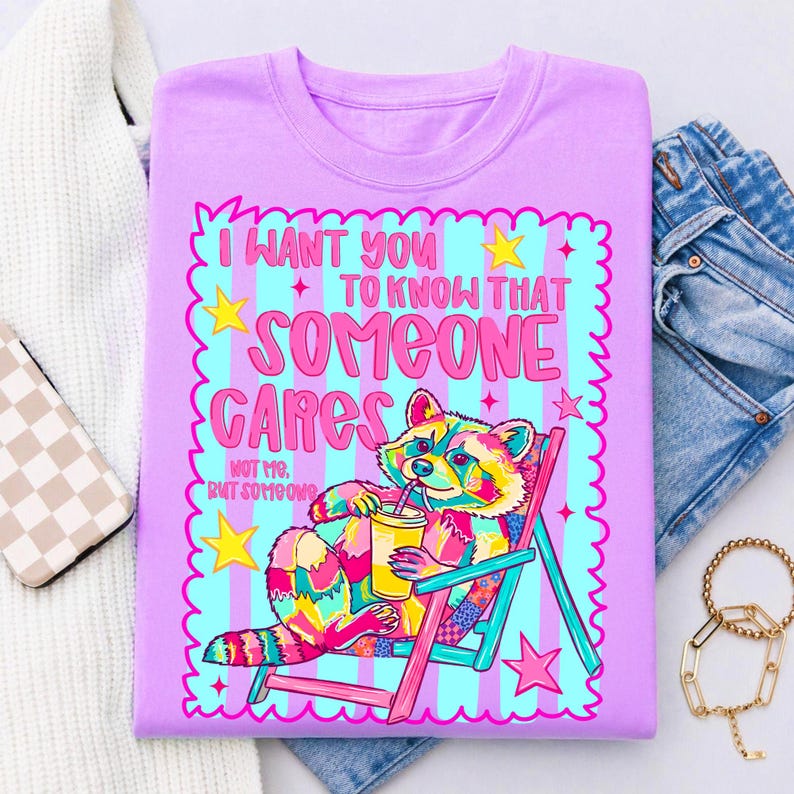 K&ouml;nnte beinhalten: Ein hellviolettes T-Shirt mit einer bunten Grafik eines Waschb&auml;ren, der in einem Strandstuhl sitzt und einen gelben Becher h&auml;lt. Der Text lautet I WANT YOU TO KNOW THAT SOMEONE CARES NOT ME, BUT SOMEONE. Das Design enth&auml;lt rosa und blaue Streifen.