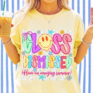 May include: A pale yellow t-shirt featuring a graphic print with the text "CLASS DISMISSED" and "Have an amazing summer". The design includes a yellow checkered smiley face, colorful letters with polka dot patterns, pink bows, and blue starfish.