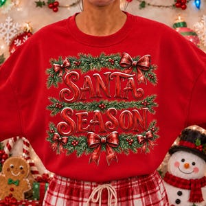 以下が含まれることがあります： 赤い文字でSANTA SEASONと書かれた鮮やかな赤色のクルーネックのスウェットシャツ。デザインは緑の松の枝、赤いベリー、そして長方形のグラフィックの四隅に配置された4つの赤のチェック柄のリボンで縁取られています。