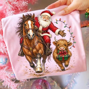 Puede incluir: Una camiseta rosa claro con una ilustraci&oacute;n gr&aacute;fica de Santa Claus montando un caballo marr&oacute;n, acompa&ntilde;ado por una peque&ntilde;a y esponjosa vaca Highland que sostiene un regalo verde con un lazo rojo. Una cadena de luces navide&ntilde;as se arquea arriba.