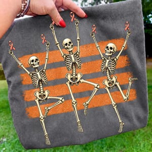 May include: A dark grey fabric item featuring a graphic print of three beige skeletons in dynamic poses. Each skeleton holds a small peach-coloured awareness ribbon above its head. Behind the skeletons are three horizontal orange brushstroke stripes.