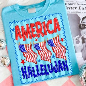 Puede incluir: Una camiseta azul claro con un estampado gr&aacute;fico que incluye el texto rojo AMERICA y el texto azul HALLELUJAH. Entre las palabras hay tres banderas estadounidenses colgadas de un tendedero, rodeadas por un borde festoneado con estrellas.