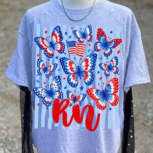 May include: A light grey t-shirt featuring a graphic print of multiple butterflies with red, white, and blue wing patterns. The design includes a small American flag, scattered red stars, light blue vertical stripes, and the red cursive text RN.