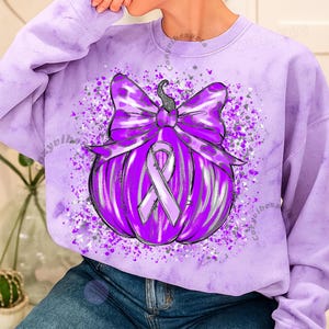 May include: A purple sweatshirt featuring a graphic of a pumpkin with a large bow and a ribbon. The pumpkin is purple with silver accents and the bow is a darker shade of purple. The ribbon is a lighter purple color.