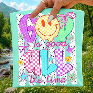 May include: A mint green t-shirt featuring a graphic design with the text "God is good all the time". The design includes a yellow checkered smiley face, colorful letters with checkered patterns, pink bows, stars, and small flowers on a dotted background.