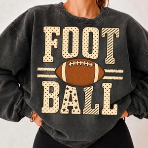 May include: A charcoal grey crewneck sweatshirt featuring the word FOOTBALL in large, cream-colored block letters. The letters have varied patterns including dots, stripes, and stars. A brown football graphic sits between the two rows of text.
