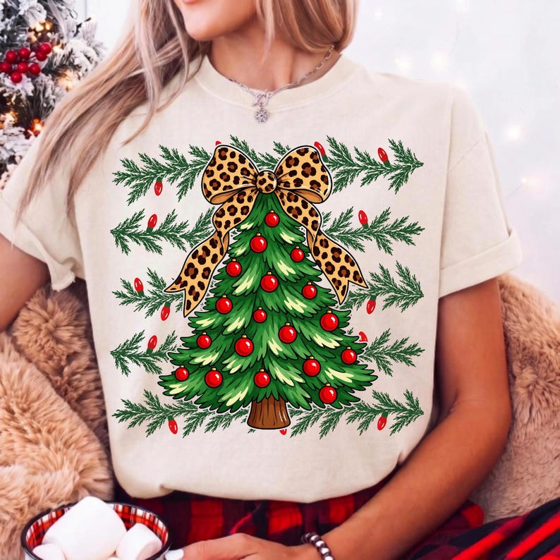 Op de afbeelding: Een cr&egrave;mekleurig T-shirt met korte mouwen en een afbeelding van een groene kerstboom versierd met rode kerstballen. De boom is bekroond met een grote strik met luipaardprint en omringd door horizontale dennentakken met rode lichtjes.