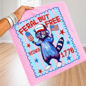May include: A pink square fabric item featuring a graphic of a raccoon wearing a red hat with the text Merica, star-patterned sunglasses, and white clogs. The raccoon holds a flag and a popsicle. Text reads FERAL BUT FREE SCINCE 1776.