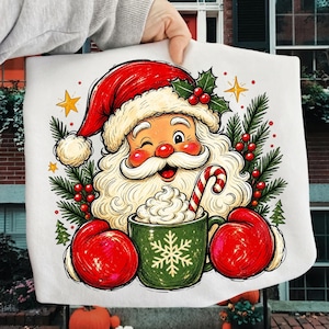 May include: A white sweatshirt featuring a graphic illustration of Santa Claus wearing a red hat and red mittens, holding a green mug with a white snowflake design, filled with whipped cream and a candy cane, surrounded by holly and pine branches.