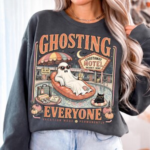 Puede incluir: Una sudadera gris carb&oacute;n de cuello redondo con un gr&aacute;fico de un fantasma con gafas de sol descansando en un flotador naranja. El dise&ntilde;o incluye un letrero de motel que dice GHOSTAWAY MOTEL, un gato negro, calabazas y el texto GHOSTING EVERYONE.