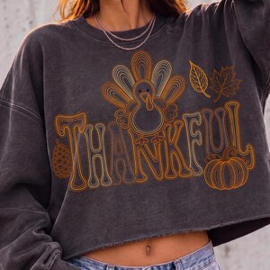 May include: A charcoal gray cropped sweatshirt featuring a graphic print of a turkey, a pinecone, a pumpkin, and two leaves. The word THANKFUL is written in large, stylized orange and yellow outline lettering across the center of the garment.