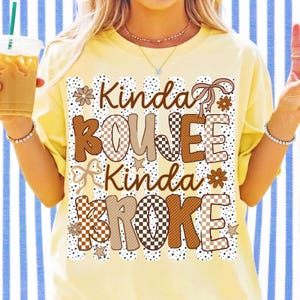 Pode incluir: Uma t-shirt amarelo-p&aacute;lido com uma estampa gr&aacute;fica de riscas verticais e pontos. O texto Kinda BOUJEE Kinda BROKE apresenta padr&otilde;es de xadrez e pontos em castanho e bege, acentuado com flores, estrelas e la&ccedil;os de fita castanhos.