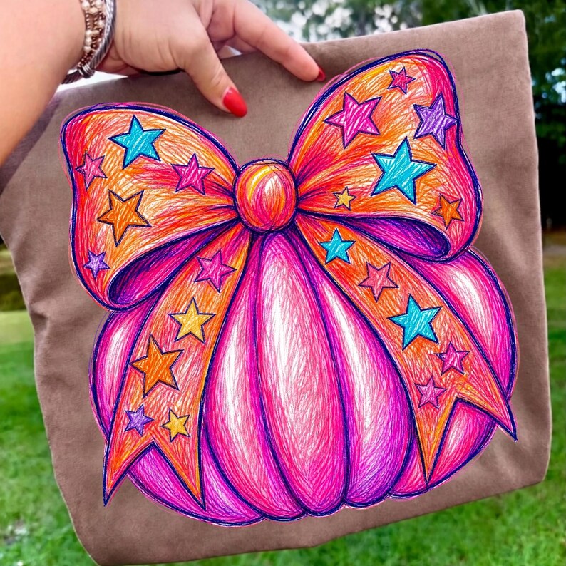 May include: A brown fabric item featuring a large, hand-drawn style graphic of a pumpkin topped with a prominent bow. The pumpkin and bow are colored in vibrant shades of pink, orange, and yellow, decorated with numerous blue, purple, and yellow stars.