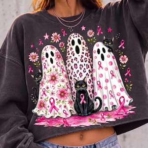 May include: A charcoal gray sweatshirt featuring a graphic of three white ghosts and three black cats. The ghosts are decorated with pink breast cancer awareness ribbons, floral patterns, leopard spots, and polka dots. Pink flowers surround the ghosts.