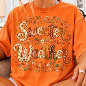 Pu&ograve; includere: Una t-shirt arancione a maniche corte con la scritta Sweater Weather in un grande carattere decorativo a motivi vari. Il design &egrave; circondato da un'illustrazione dettagliata di varie foglie autunnali, piccoli fiori e viticci.