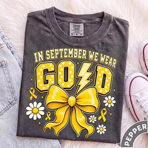 May include: A charcoal gray t-shirt featuring the text IN SEPTEMBER WE WEAR GOLD in yellow block letters with white polka dots. The design includes a large yellow bow, a lightning bolt, small daisies, and yellow awareness ribbons.