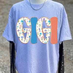 May include: A light gray short-sleeve t-shirt featuring the word GIGI in large letters. The G and I characters have a cream background with a colorful floral pattern, while the other G and I characters feature blue and orange polka dot patterns.