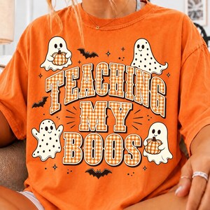May include: An orange short-sleeve t-shirt featuring a graphic print with the text TEACHING MY BOOS in a white and orange checkered pattern. The design includes four white ghosts, two of which are holding orange pumpkins, and three black bats.