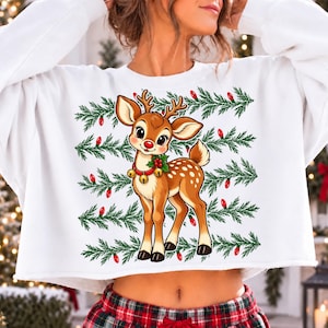 May include: A white long-sleeve sweatshirt featuring a central graphic of a brown fawn with large eyes, a red nose, and a collar with gold bells and holly. The background of the print consists of horizontal green pine branches and red light bulbs.