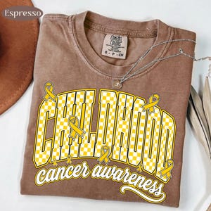Puede incluir: Una camiseta color espresso con un gr&aacute;fico que presenta el texto CHILDHOOD en un patr&oacute;n de cuadros amarillos y blancos, y cancer awareness en letra cursiva blanca. Varios lazos amarillos est&aacute;n integrados en el dise&ntilde;o.