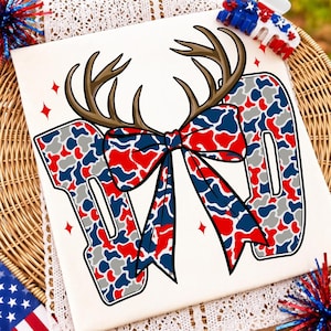May include: A white t-shirt featuring a graphic of the number 100 in a red, white, and blue camouflage pattern. The design includes a large brown deer antler set above the numbers, with a matching red, white, and blue patterned bow tied at the centre.