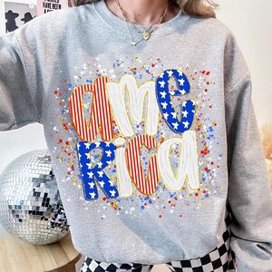 K&ouml;nnte beinhalten: Ein graues Sweatshirt mit dem Wort "America" in einem patriotischen Design. Die Buchstaben sind rot-wei&szlig; gestreift und blau mit wei&szlig;en Sternen. Das Design enth&auml;lt rote, wei&szlig;e und blaue Spritzer.