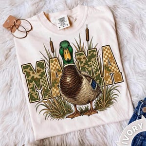 May include: A cream-colored t-shirt featuring a graphic of a mallard duck standing in tall grass. The word MAMA is printed in large, block letters with a green camouflage and tan checkered pattern. The shirt includes a Comfort Colors brand tag.