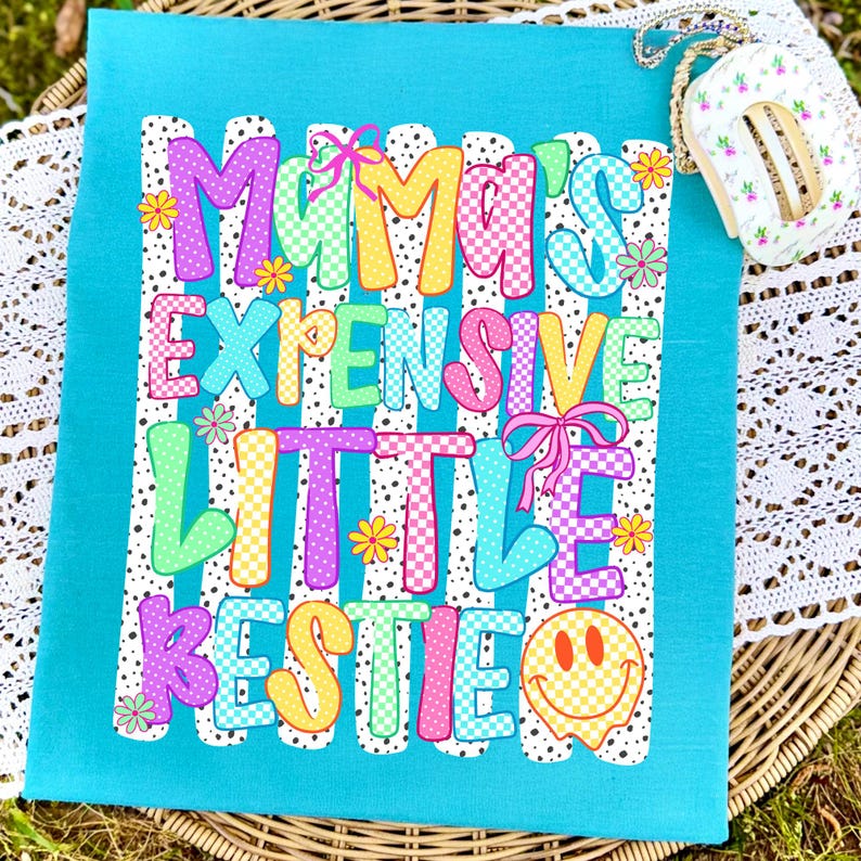 May include: A bright turquoise t-shirt featuring a graphic print with the text "MAMA'S EXPENSIVE LITTLE BESTIE" in colorful, patterned bubble letters. The design includes pink bows, small flowers, and a yellow smiley face on a white striped background.