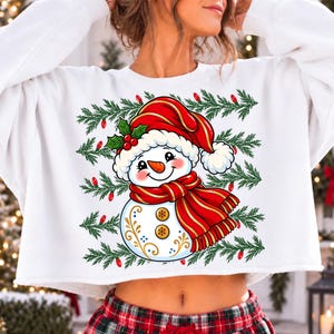 May include: A white long-sleeve cropped sweatshirt featuring a graphic of a smiling snowman wearing a red hat and scarf, accented with holly leaves and berries. The design is surrounded by horizontal rows of green pine branches and small red lights.
