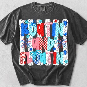 Op de afbeelding: Een antracietgrijs t-shirt met korte mouwen en een grafische print met de tekst "BOATIN' AND FLOATIN'". Het ontwerp bevat verticale witte strepen met zwarte stippen, rood, blauw en roze geblokte letters, blauwe strikken en vuurwerkillustraties.