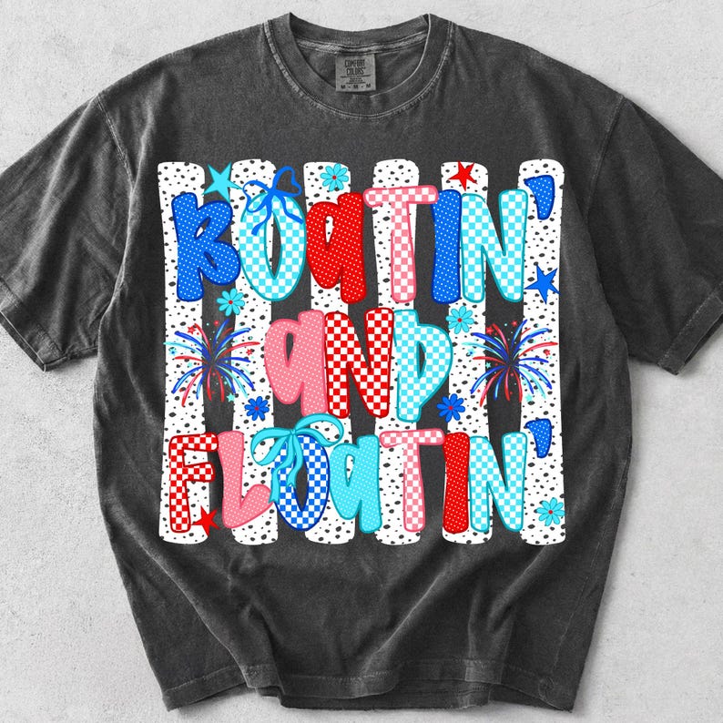Op de afbeelding: Een antracietgrijs t-shirt met korte mouwen en een grafische print met de tekst "BOATIN' AND FLOATIN'". Het ontwerp bevat verticale witte strepen met zwarte stippen, rood, blauw en roze geblokte letters, blauwe strikken en vuurwerkillustraties.