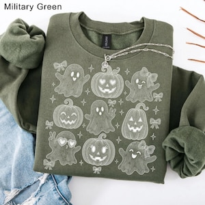 Puede incluir: Una sudadera de cuello redondo color verde militar con un gr&aacute;fico de l&iacute;neas blancas que muestra varios fantasmas y calabazas. Los fantasmas tienen diferentes expresiones y accesorios como lazos y gafas en forma de coraz&oacute;n.