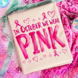 May include: A beige t-shirt featuring the hot pink text IN OCTOBER WE WEAR PINK in a brushstroke style. The design includes small pink heart icons, ribbon symbols, and four-pointed star accents arranged around the bold, centred lettering.