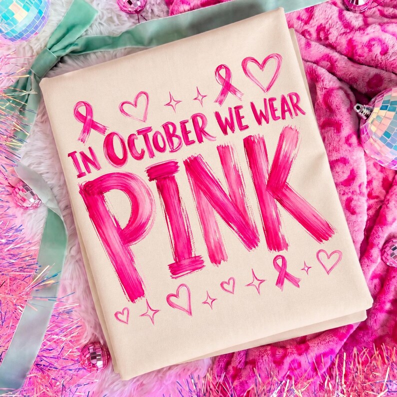 May include: A beige t-shirt featuring the hot pink text IN OCTOBER WE WEAR PINK in a brushstroke style. The design includes small pink heart icons, ribbon symbols, and four-pointed star accents arranged around the bold, centred lettering.