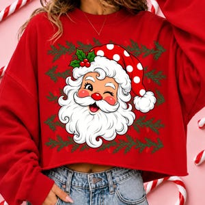 Puede incluir: Una sudadera roja de manga larga con un gr&aacute;fico de un Santa Claus gui&ntilde;ando un ojo, que lleva un gorro rojo con lunares blancos y hojas de acebo. El dise&ntilde;o est&aacute; enmarcado por un patr&oacute;n repetitivo de ramas de pino verdes en la parte frontal.