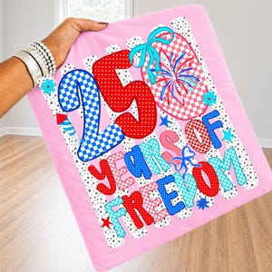 May include: A pink fabric square featuring the text 25 YEARS OF FREEDOM in red, blue, and light blue checkered and polka dot patterns. The design includes a firework graphic, blue ribbons, and small stars against a white background with black dots.