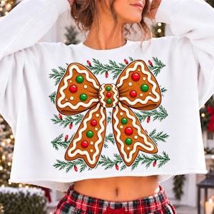 Op de afbeelding: Een wit kort sweatshirt met een grote afbeelding van een peperkoekstrik, versierd met wit glazuur en rode en groene snoepjes. Het ontwerp is omlijst door horizontale rijen groene dennentakken met daartussen kleine rode kerstlichtjes.