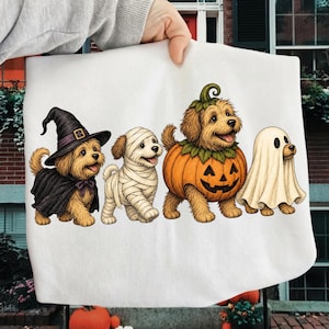 Puede incluir: Una superficie de tela blanca con una ilustraci&oacute;n gr&aacute;fica de cuatro perros peque&ntilde;os con disfraces de Halloween. De izquierda a derecha, un perro lleva un sombrero de bruja y capa negra, un perro envuelto en vendas blancas, un perro dentro de una calabaza y un perro de fantasma.