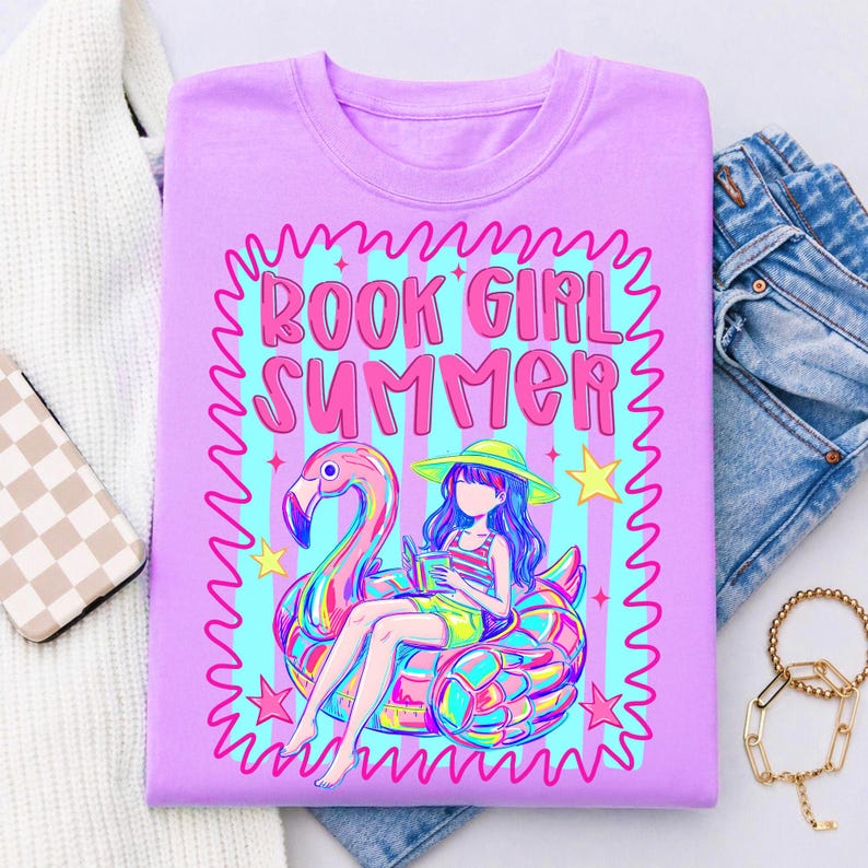 May include: A light purple t-shirt featuring a graphic print with the text "BOOK GIRL SUMMER" in pink. The design includes a person with long hair wearing a hat, sitting on a colourful flamingo pool float while reading a book, surrounded by stars.