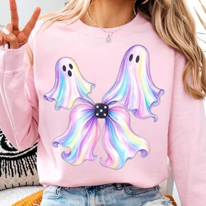 May include: A light pink crewneck sweatshirt featuring a graphic of two iridescent, pastel-coloured ghosts with flowing, wavy edges. The ghosts are positioned around a central black die with white dots, creating a bow-like shape on the chest.