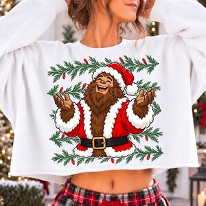 Puede incluir: Una sudadera blanca de manga larga y corte corto con un gr&aacute;fico de un personaje Bigfoot marr&oacute;n que viste un traje de Pap&aacute; Noel rojo con ribete de piel blanca, un cintur&oacute;n negro con hebilla dorada y un gorro rojo, rodeado de ramas de pino verdes y luces rojas.