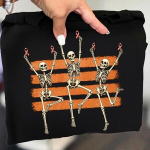 May include: A black sweatshirt featuring a graphic print of three white skeletons in dynamic dancing poses. Each skeleton holds a small pink awareness ribbon above its head. The design is set against three horizontal, distressed orange brush strokes.