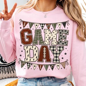 May include: A light pink crewneck sweatshirt featuring a graphic print with the text GAME DAY in large block letters. The letters are filled with various patterns including footballs, stars, and plaid. A bunting banner with football motifs frames the text.
