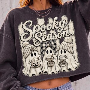 May include: A charcoal gray crewneck sweatshirt featuring a graphic print with the text Spooky Season. The design displays three white ghosts wearing knit hats, one with a buffalo plaid bow, holding a pumpkin mug and two coffee cups.