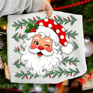 Puede incluir: Una camiseta blanca con un gr&aacute;fico de un Santa Claus gui&ntilde;ando un ojo con un gorro rojo de lunares blancos, hojas de acebo verdes y bayas rojas, rodeado por un patr&oacute;n repetitivo de ramas de pino verdes y peque&ntilde;as bombillas navide&ntilde;as rojas.
