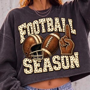 Puede incluir: Una sudadera corta de color gris carb&oacute;n con un estampado gr&aacute;fico que muestra el texto FOOTBALL SEASON en una fuente de color crema con lunares negros. El dise&ntilde;o incluye un casco de f&uacute;tbol americano marr&oacute;n, un bal&oacute;n y un dedo de espuma marr&oacute;n.