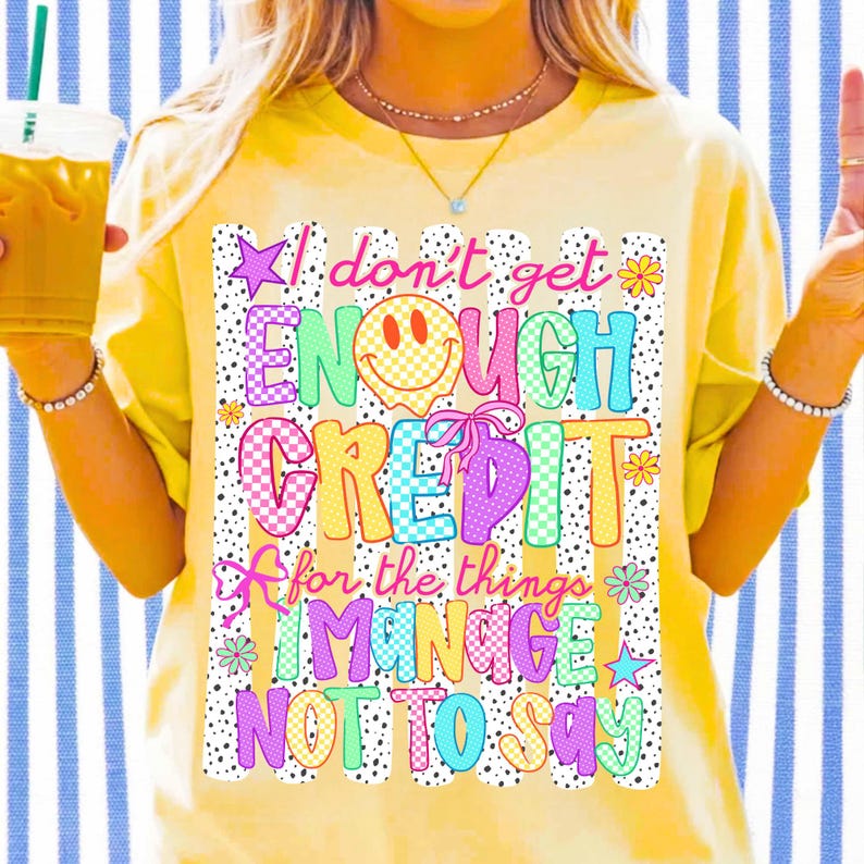 Pode incluir: Uma t-shirt de manga curta em amarelo p&aacute;lido com uma impress&atilde;o gr&aacute;fica colorida com o texto I don't get ENOUGH CREDIT for the things I MANAGE NOT TO SAY. O design inclui uma cara sorridente amarela, la&ccedil;os cor-de-rosa, estrelas e flores.
