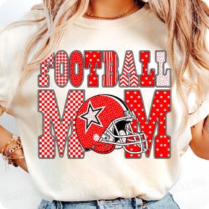 May include: Cream-colored t-shirt with the words "FOOTBALL MOM" in red, patterned text. A red football helmet with a white star is featured in the design. The shirt is a casual, sporty style.
