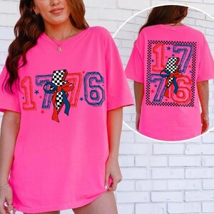 Op de afbeelding: Een felroze T-shirt met korte mouwen en een afbeelding van het getal 1776 met een geblokte bliksemschicht en een rode en blauwe strik. Het ontwerp bevat kleine blauwe en rode sterren, met een grotere versie van de afbeelding op de achterkant.